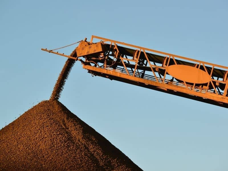 Orange industrial conveyor pours gravel into a large mound under a clear blue sky, outdoors.